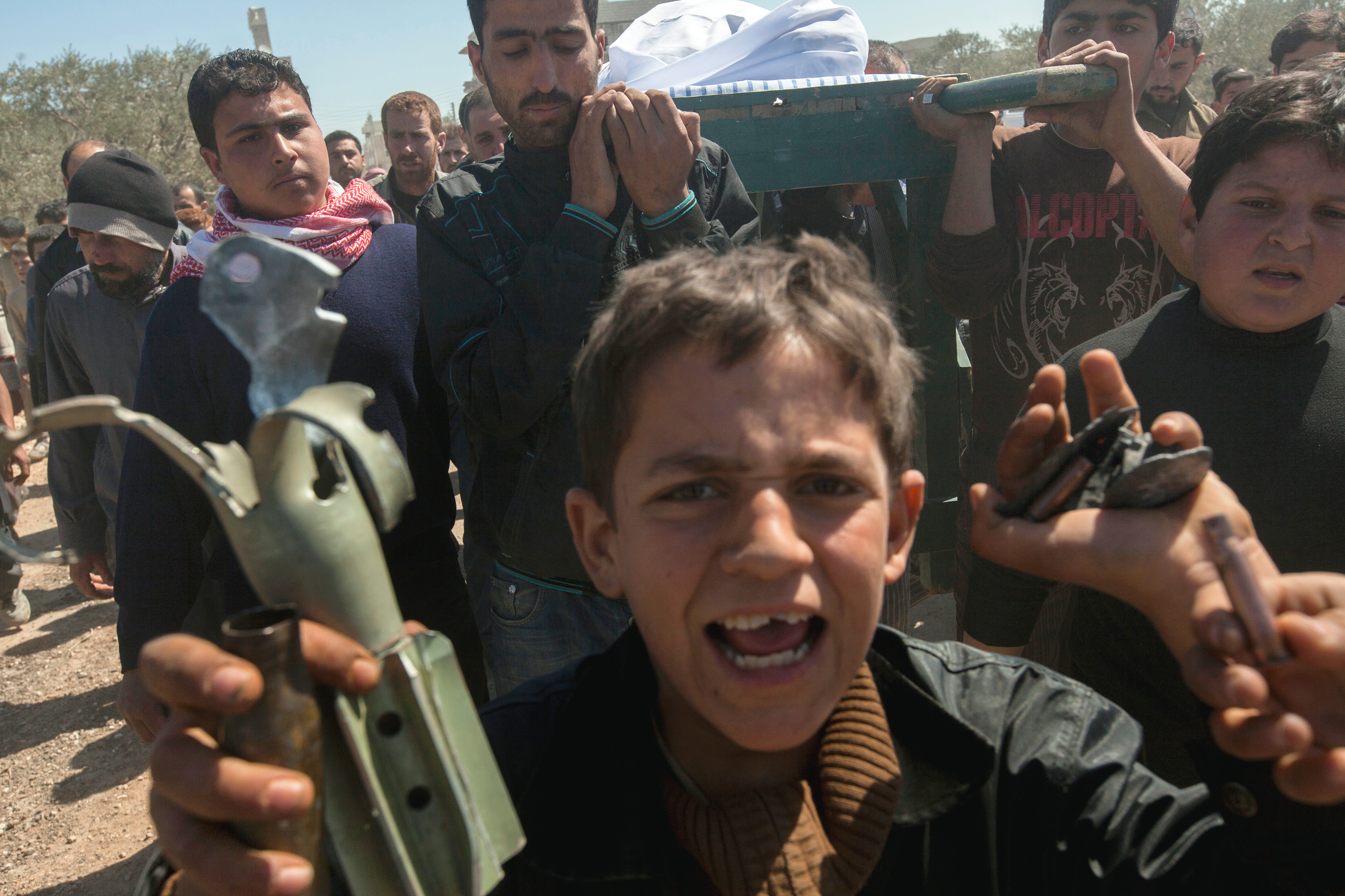 A grieving boy holds a broken weapon fragment while shouting, as a group of people behind him carry a body wrapped in a white shroud during a funeral procession. The crowd appears solemn and distressed.