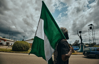 Afrian man holding a Nigerian flag walking down a street with houses and a cloudy sky in the background