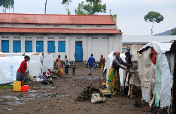 Muddy walkway between rows of white tents in front of a large building with blue windows