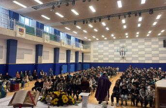 A large group of children and adults sit in a gymnasium, facing a speaker in a purple gown at the front. Sunflowers and an open book are visible in the foreground. The gym has blue and white walls and high windows.