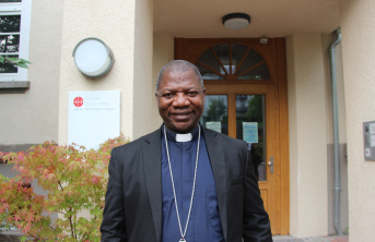 A man wearing clerical clothing and a cross necklace stands smiling in front of a building entrance with a wooden door and a sign on the wall.