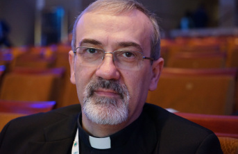 A middle-aged man with glasses, grey hair, and a short beard, wearing a clerical collar and dark clothing, sits in an auditorium with empty brown seats in the background