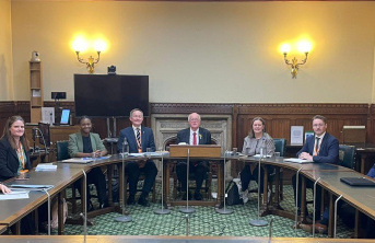 A group of eight people, four men and four women, sit around a curved table in a formal meeting room with wood panelling, chandeliers, and a fireplace. The atmosphere is professional and official.