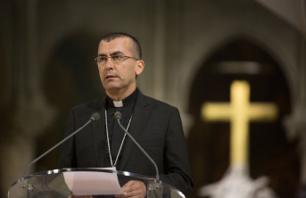 Priest standing in front of a microphone with a cross blurred in the background