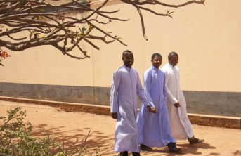 3 Nigerian seminarians walking across sand with a tree and bush in the foreground