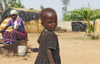A young Nigerian girl in a dark dress standing as her mother looks on, seated while cooking from a metal pot.