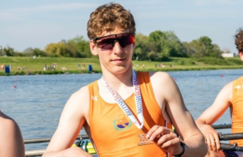 Teenage boy wearing sunglasses holding a medal in a rowing boat with river behind him