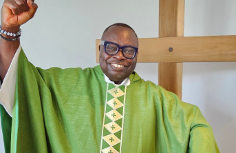 A priest in green standing in front of a cross, smiling with arm raised in the air in celebration