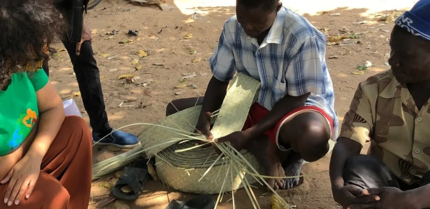 Two African men kneeling and demonstrating their craft to a woman in a green top
