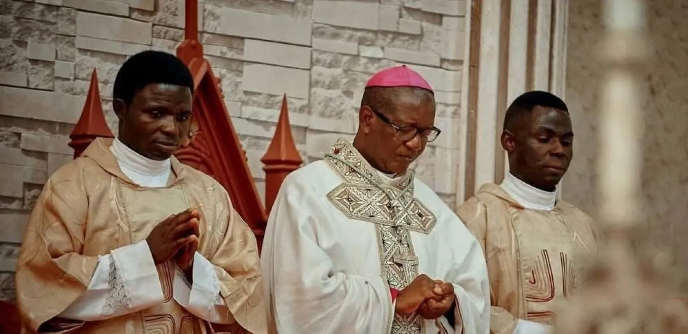 Three Nigerian men including a Bishop in the middle dressed in religious clothing bowing their heads in prayer