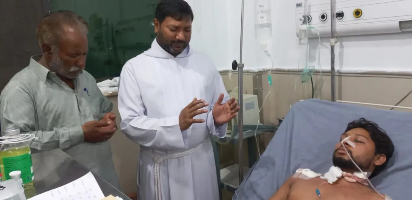 A Pakistani Father and a man standing over another man in a hospital bed giving prayer
