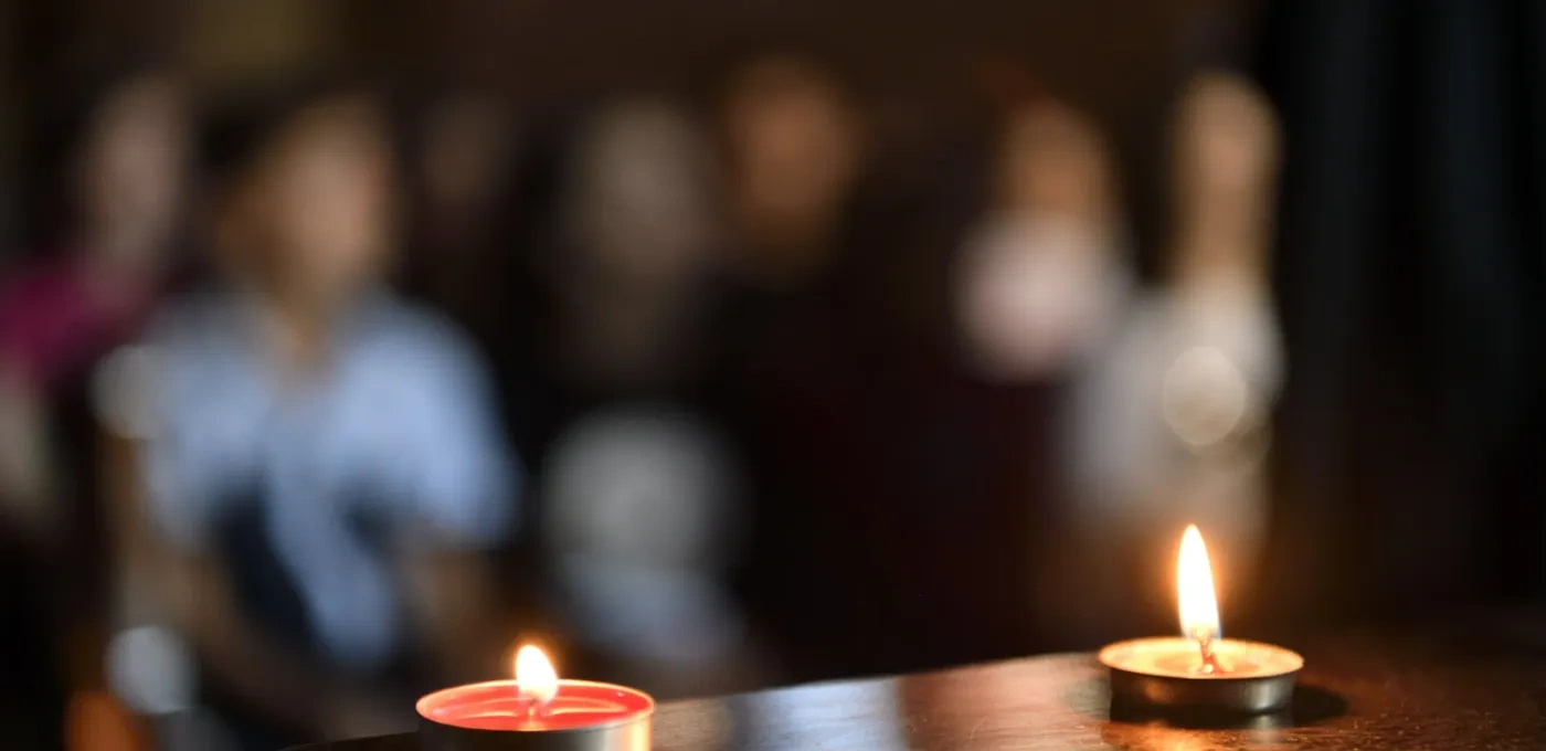 Two lit candles on a table with a blurred background of people sitting in prayer