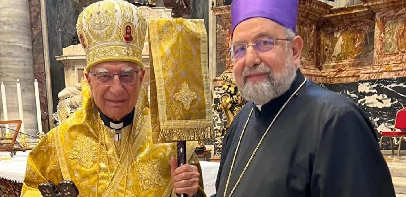 Two elderly men standing together in front of an alter. Man on left wearing gold religious clothing