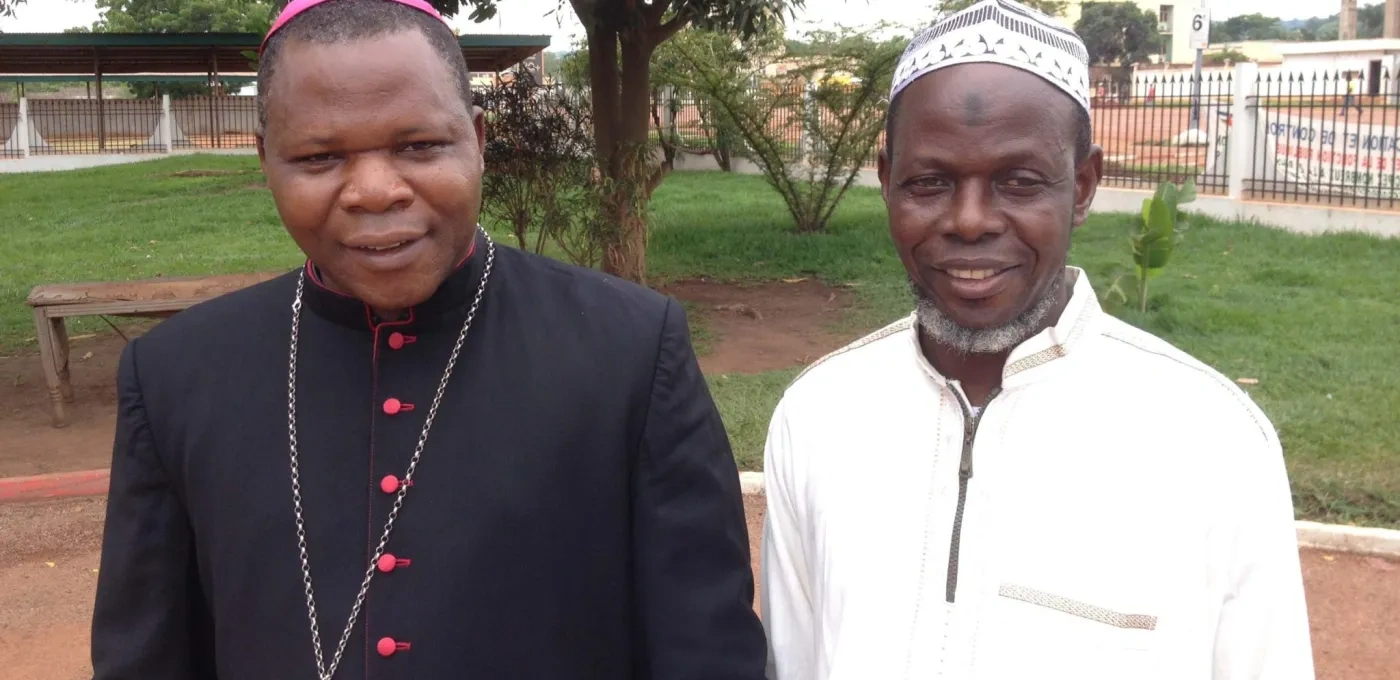Two African men standing besides each other with one wearing white and the other in Cardinal's black clothing