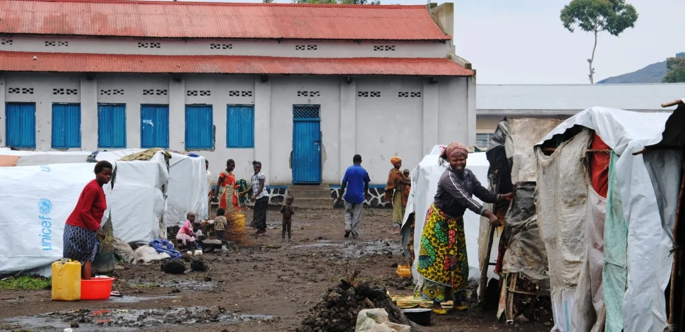 Muddy walkway between rows of white tents in front of a large building with blue windows