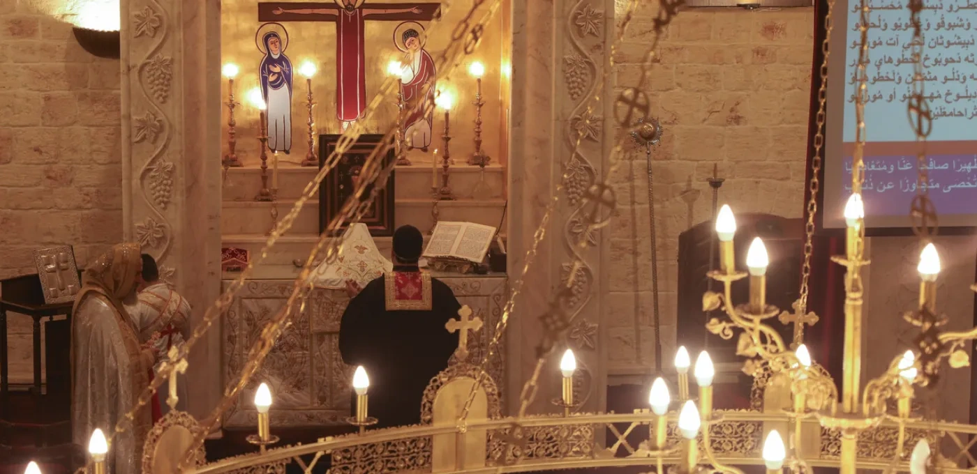 Interior of a church with lit candles in the foreground and a priest praying to the cross in the background