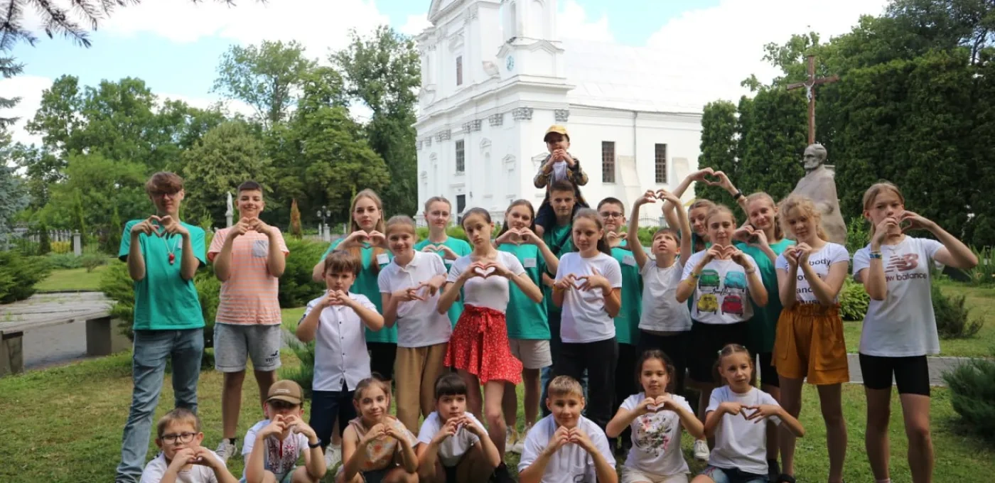 A group of young children, with some sitting on grass and all making a heart shape with their hands