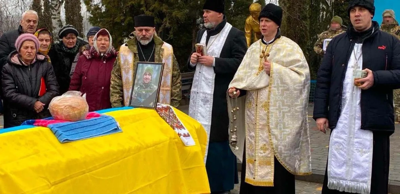 A Father gathered with a group of adult men and women at a funeral overlooking a coffin draped with a Ukrainian flag