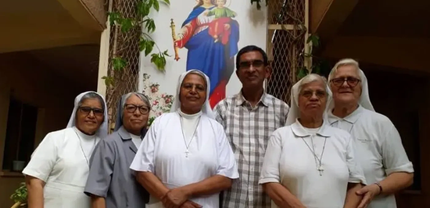 A priest and Sisters standing in front of image of Jesus and Mary
