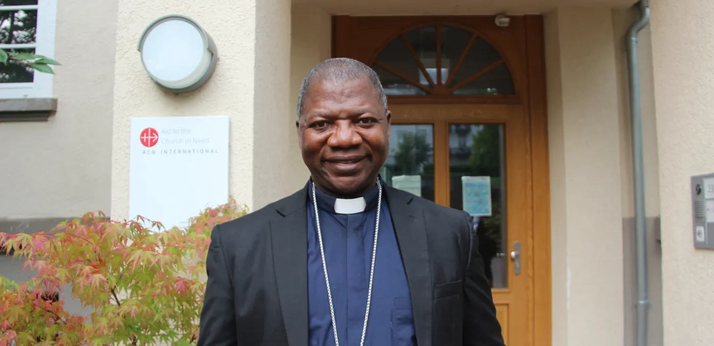 A man wearing clerical clothing and a cross necklace stands smiling in front of a building entrance with a wooden door and a sign on the wall.