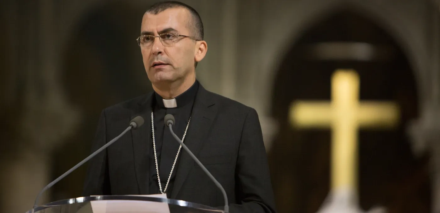 Priest standing in front of a microphone with a cross blurred in the background
