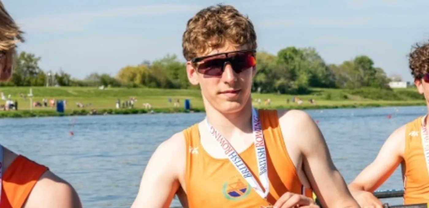 Teenage boy wearing sunglasses holding a medal in a rowing boat with river behind him