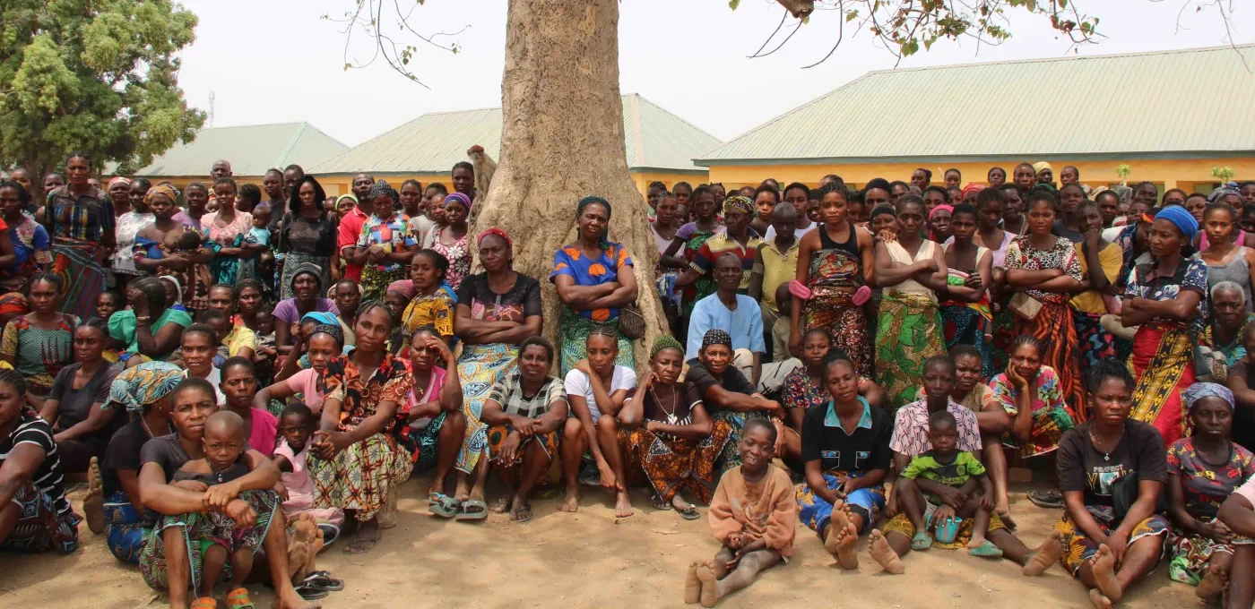A large group of Nigerian people that have been displaced sitting and standing under a tree
