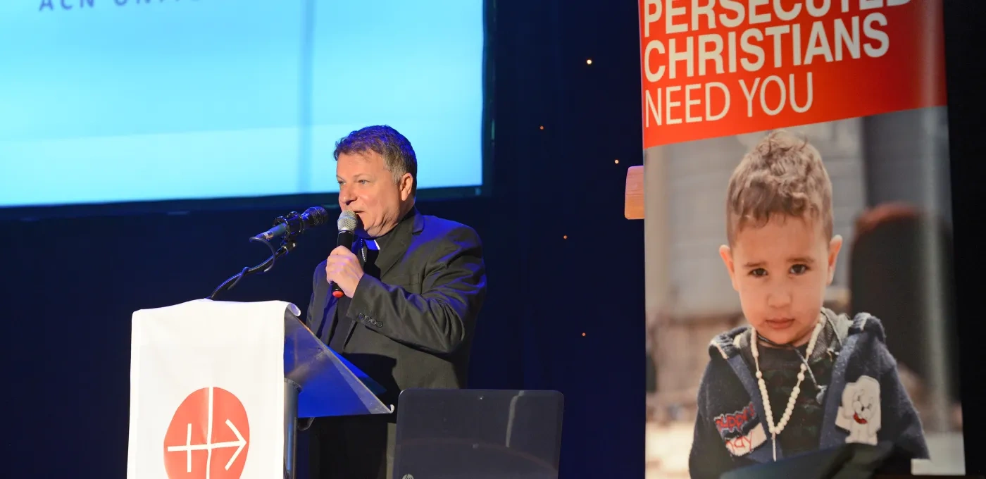 A priest standing in front of podium with an ACN charity banner behind him