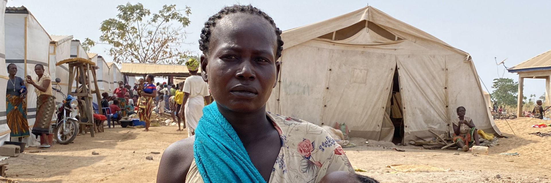 A Nigerian mother her child standing in front of a tent and group of people in the background