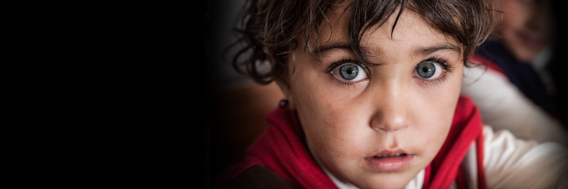 A young child with wide green eyes and tousled brown hair looks directly at the camera, wearing a red and white garment.