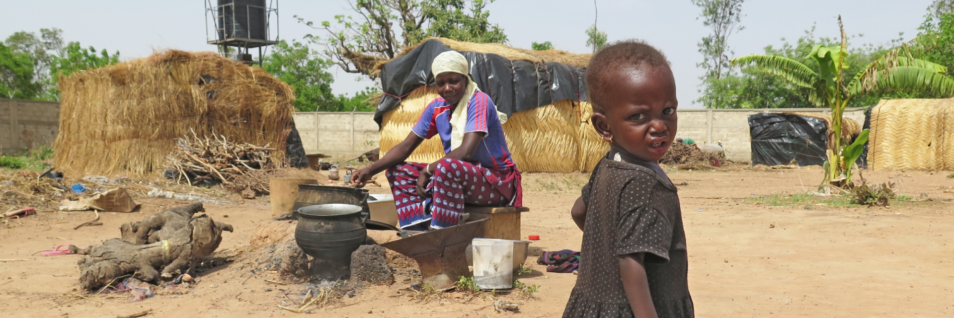 A young Nigerian girl in a dark dress standing as her mother looks on, seated while cooking from a metal pot.