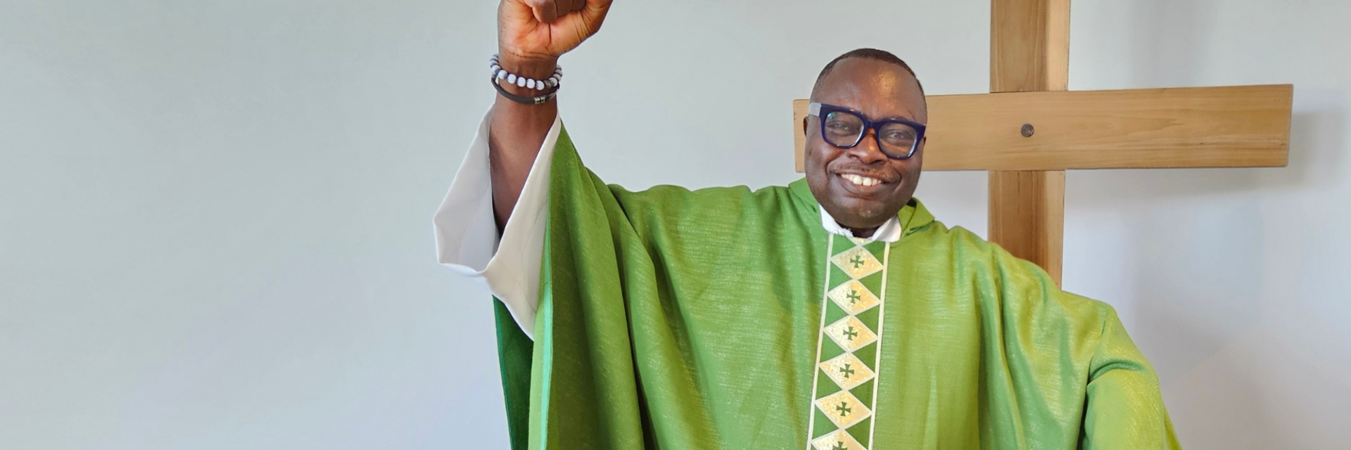 A priest in green standing in front of a cross, smiling with arm raised in the air in celebration