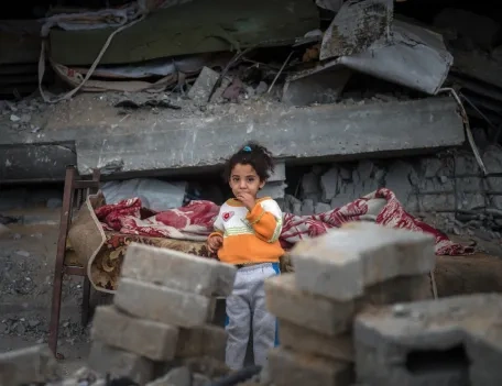A young child stands in front of a damaged building and scattered rubble, wearing a bright orange and white jumper, with a red patterned blanket on a bed behind them. The scene suggests destruction and hardship.