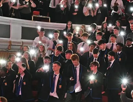 School pupils standing together at a concert hall with their flashlights lit on their mobile phones.