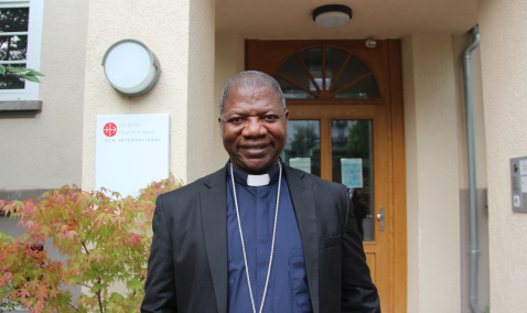 A man wearing clerical clothing and a cross necklace stands smiling in front of a building entrance with a wooden door and a sign on the wall.