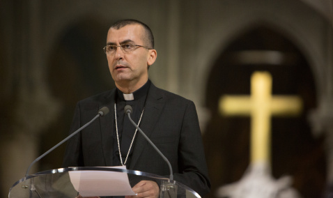 Priest standing in front of a microphone with a cross blurred in the background