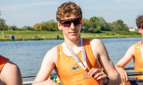 Teenage boy wearing sunglasses holding a medal in a rowing boat with river behind him