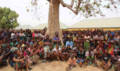 A large group of Nigerian people that have been displaced sitting and standing under a tree