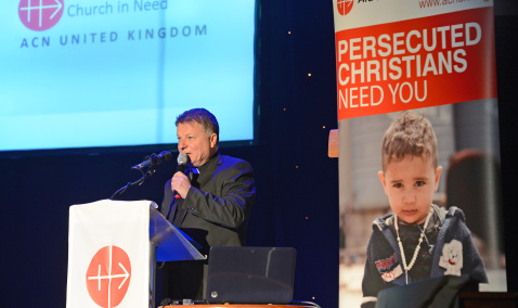 A priest standing in front of podium with an ACN charity banner behind him