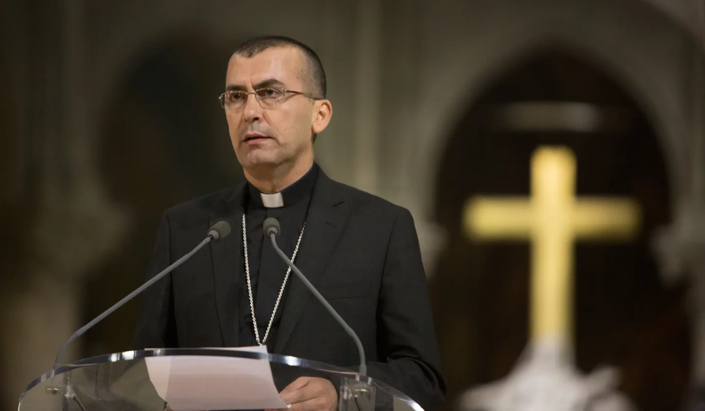 Priest standing in front of a microphone with a cross blurred in the background