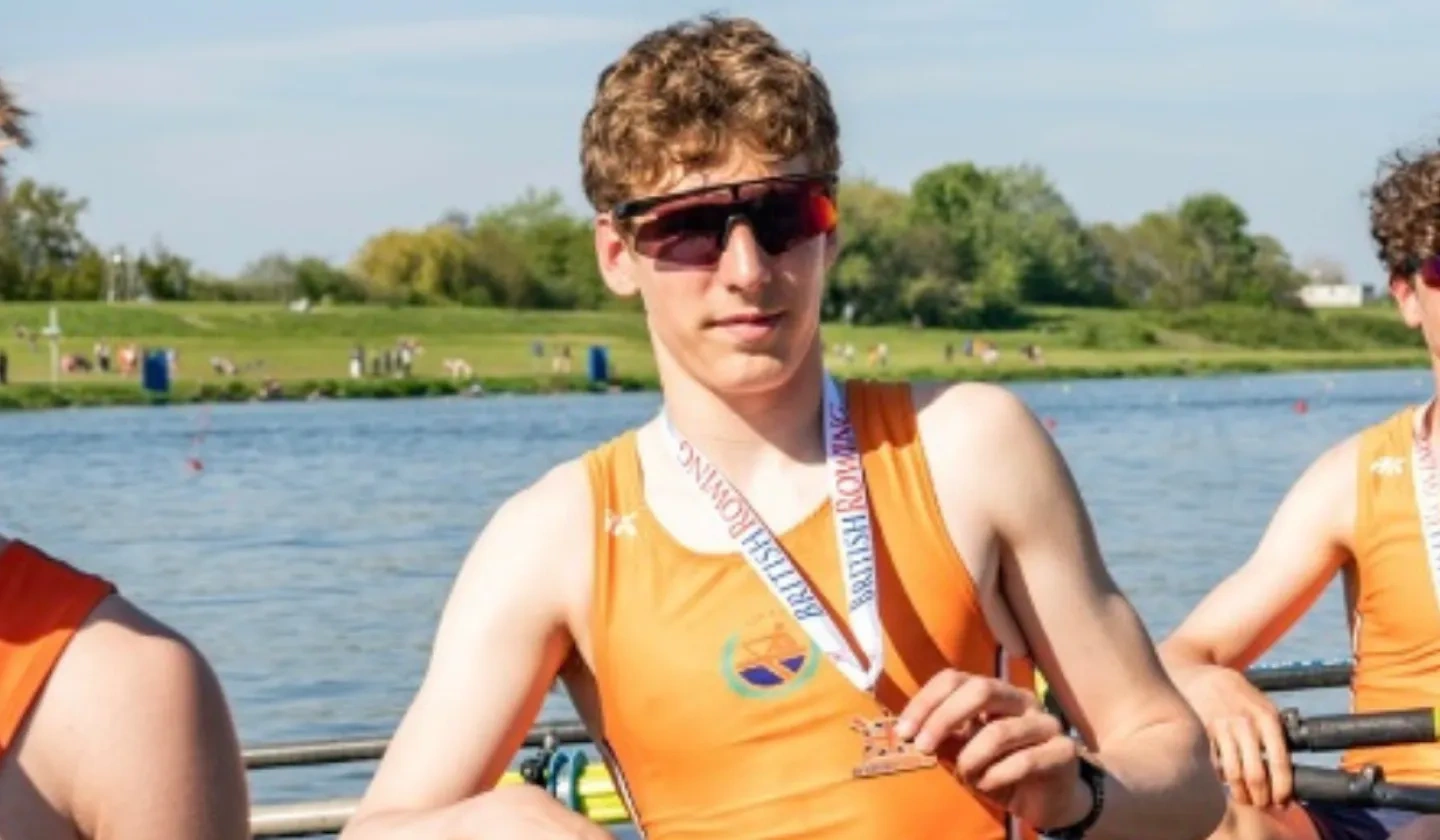 Teenage boy wearing sunglasses holding a medal in a rowing boat with river behind him