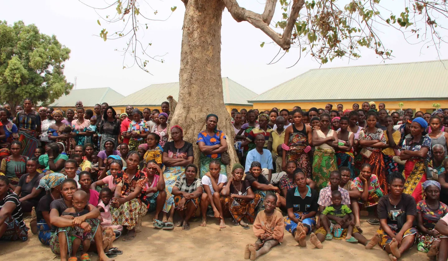 A large group of Nigerian people that have been displaced sitting and standing under a tree