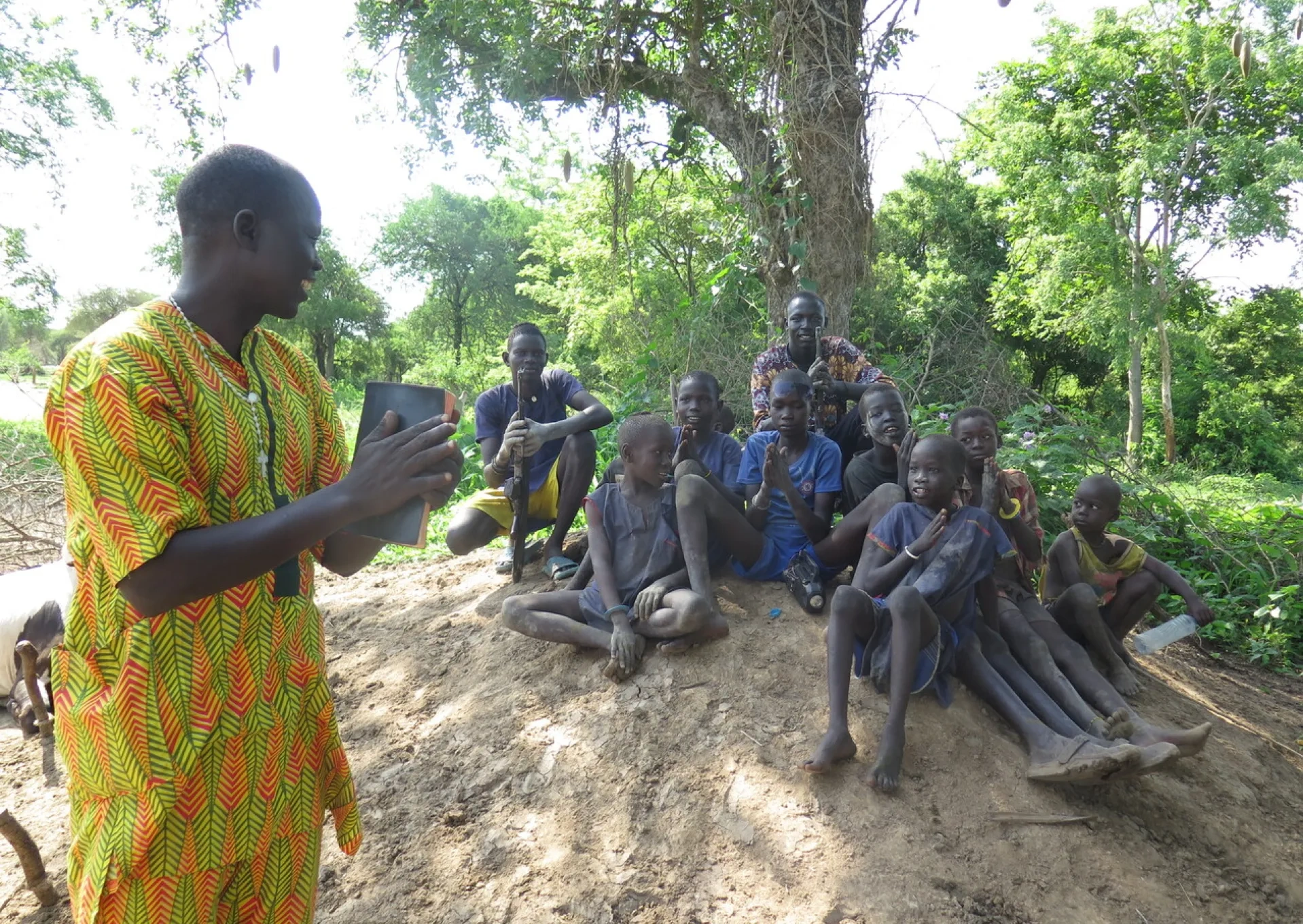 A missionary standing holding a bible talking to a group of boys sitting on a mound under a tree