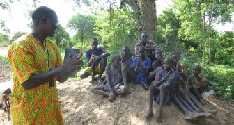 A missionary standing holding a bible talking to a group of boys sitting on a mound under a tree