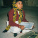 A young child sits on the floor, holding a slate and chalk, with a school bag nearby.