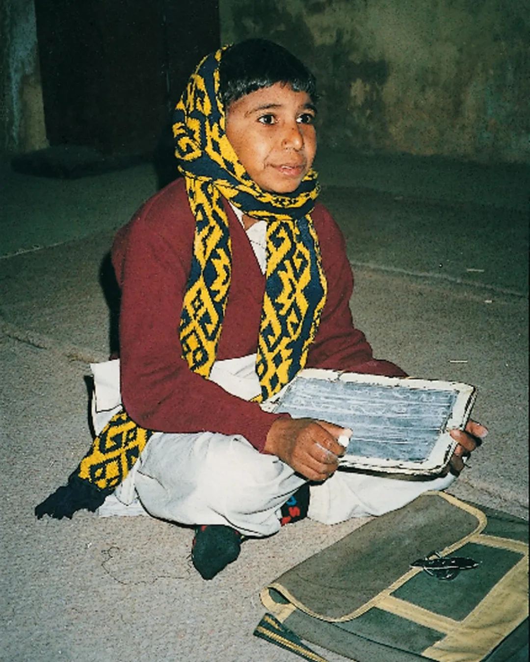 A young child sits on the floor, holding a slate and chalk, with a school bag nearby.